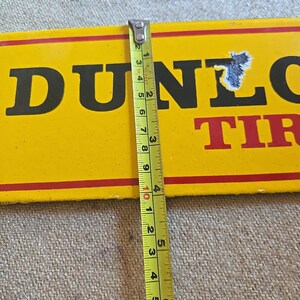K&ouml;nnte beinhalten: Ein gelbes Dunlop Tires-Schild mit schwarzer Schrift und roten Streifen. Das Schild wird mit einem Ma&szlig;band gemessen, das eine L&auml;nge von etwa 25 cm anzeigt. Das Schild hat ein abgenutztes Aussehen.