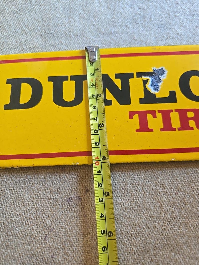 K&ouml;nnte beinhalten: Ein gelbes Dunlop Tires-Schild mit schwarzer Schrift und roten Streifen. Das Schild wird mit einem Ma&szlig;band gemessen, das eine L&auml;nge von etwa 25 cm anzeigt. Das Schild hat ein abgenutztes Aussehen.