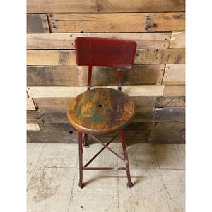 May include: A stool with a round, weathered wooden seat featuring a central hole and a red metal frame. The backrest is a rectangular red metal panel attached to the frame. The legs are connected by a cross-brace structure for stability.
