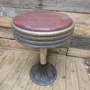 May include: Vintage diner-style stool with a round, burgundy seat and a chrome-plated, ribbed base. The base has a silver-colored, adjustable column and a black, circular footrest. The stool is designed for seating.