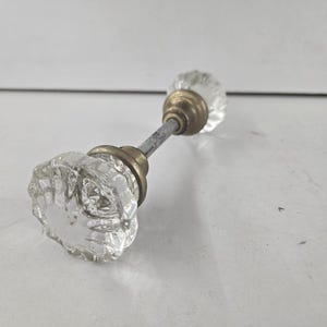 May include: A door knob set featuring two clear, faceted glass knobs with a starburst pattern. Each knob is attached to a circular, tarnished brass-toned metal base, and the two knobs are connected by a central, thin, silver-toned metal spindle.
