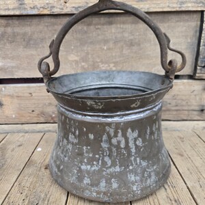 May include: A weathered, round metal bucket with a wide, flared rim and a sturdy, arched handle attached to the sides. The exterior surface features a mottled, tarnished finish with visible patches of silver and dark brown oxidation throughout.