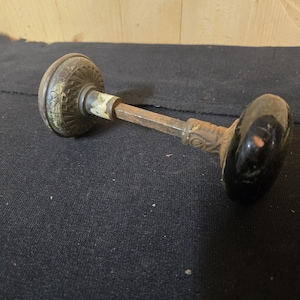 May include: Antique door knob with a decorative, aged brass finish on one end and a black, oval-shaped knob on the other. The connecting rod is metal with signs of wear. The knob is likely from a vintage or historical home.
