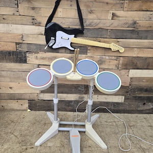Può includere: Un controller per chitarra elettrica bianco e nero con tracolla nera e una batteria elettronica a quattro pezzi con pedale e due bacchette. I pad della batteria presentano bordi circolari rossi, gialli, blu e verdi su un supporto grigio chiaro.