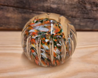 May include: A clear glass paperweight with a swirl of colorful glass inside. The glass is a mix of white, orange, green, and black.