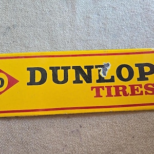 K&ouml;nnte beinhalten: Ein rechteckiges gelbes Dunlop Tires-Schild mit rotem Rand. Das Schild zeigt das Dunlop-Logo, einen roten Pfeil mit einem wei&szlig;en "D" darin und die Worte "DUNLOP TIRES" in Schwarz und Rot.