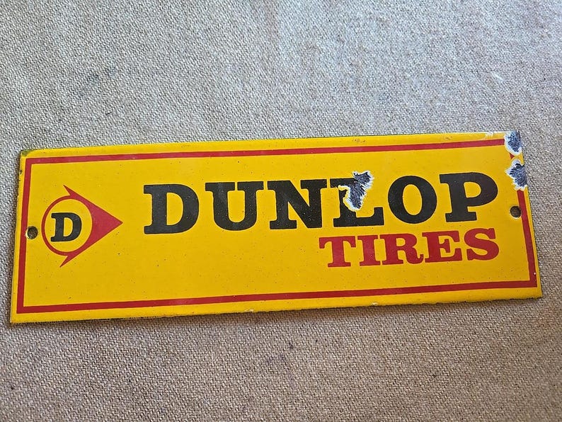 K&ouml;nnte beinhalten: Ein rechteckiges gelbes Dunlop Tires-Schild mit rotem Rand. Das Schild zeigt das Dunlop-Logo, einen roten Pfeil mit einem wei&szlig;en "D" darin und die Worte "DUNLOP TIRES" in Schwarz und Rot.