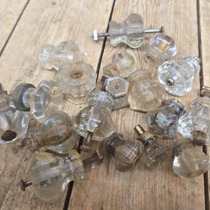 May include: A collection of clear glass cabinet knobs featuring a faceted, multi-sided design. Each knob includes a metal screw or bolt attachment, with some showing signs of age, oxidation, and varying levels of transparency and wear.