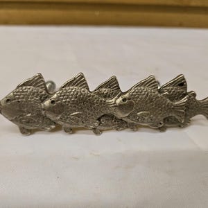 May include: A silver-toned metal cabinet pull featuring a row of five fish. Each fish has detailed scales and fins, creating a textured appearance. The pull is designed to be mounted on a cabinet or drawer.