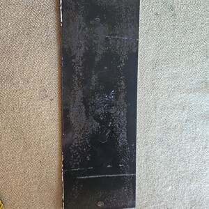 K&ouml;nnte beinhalten: Ein rechteckiges schwarzes Metallschild mit zwei L&ouml;chern oben und einem unten. Das Schild hat ein abgenutztes, verwittertes Aussehen mit wei&szlig;er Farbe an den R&auml;ndern.