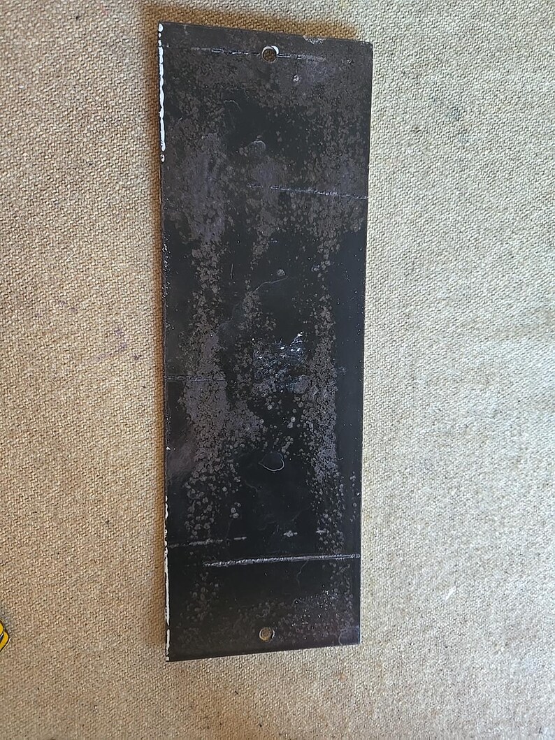 K&ouml;nnte beinhalten: Ein rechteckiges schwarzes Metallschild mit zwei L&ouml;chern oben und einem unten. Das Schild hat ein abgenutztes, verwittertes Aussehen mit wei&szlig;er Farbe an den R&auml;ndern.