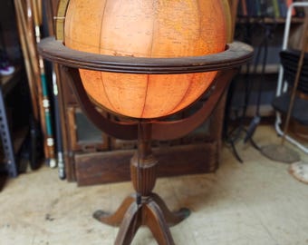 May include: An antique illuminated globe on a wooden stand. The globe is a warm orange color, with detailed cartography. The stand has a tripod base and a dark brown finish. The globe is lit from within, creating a soft glow.