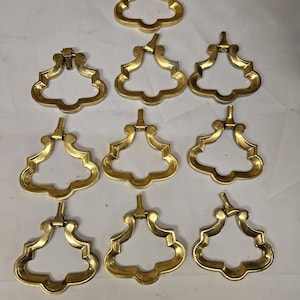 May include: Nine ornate, gold-colored drawer pulls arranged on a white surface. Each pull has a decorative, openwork design with a central handle and a hinged top for attachment. The pulls are likely for furniture.