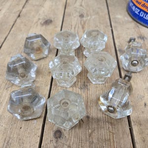 May include: A collection of ten clear glass cabinet knobs featuring a faceted hexagonal design. Each knob has a central metal mounting point and a transparent, multi-sided shape that reflects light, arranged in a group on a wooden surface.
