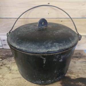 May include: A black cast iron pot with a matching lid and a wire bail handle. The pot features a weathered, matte finish with visible signs of wear and texture across the surface. A small, upright tab with a hole is centered on the lid.