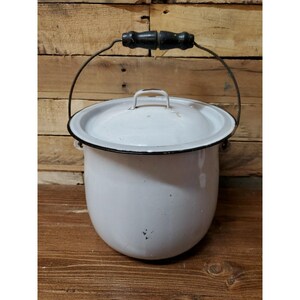 May include: A white enamel pot with a matching lid and a black rim. The pot features a metal swing handle with a black wooden grip. The exterior surface is smooth with minor dark spots and signs of wear, showing a classic cylindrical shape.