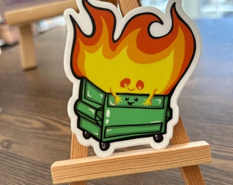 May include: A green dumpster with a smiling face is on fire. The flames are yellow and orange and the dumpster has wheels.