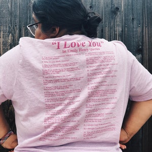 May include: A pink t-shirt with a quote from Emily Henry that reads "I Love You" in pink lettering. The quote is a list of 34 reasons why someone might love another person.