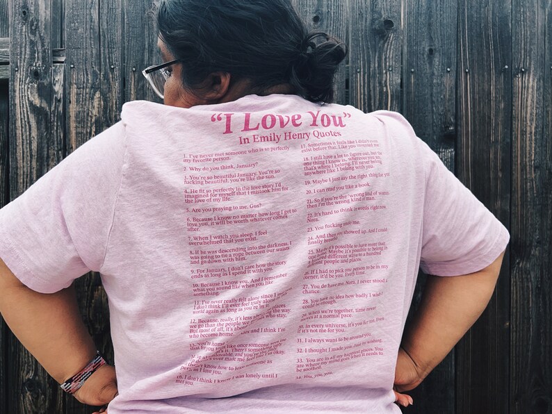 May include: A pink t-shirt with a quote from Emily Henry that reads "I Love You" in pink lettering. The quote is a list of 34 reasons why someone might love another person.