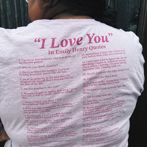 May include: A pink t-shirt with a quote from the book "I Love You" by Emily Henry. The quote is printed in a dark pink font and includes a list of 32 quotes from the book.