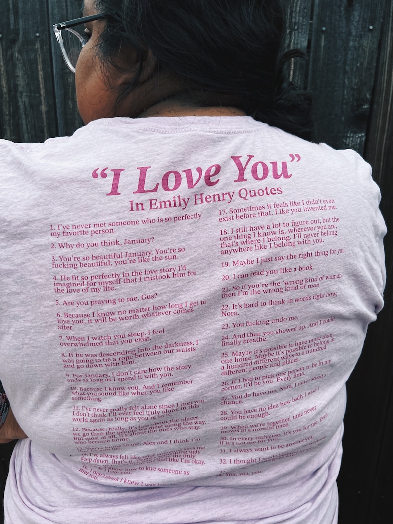 May include: A pink t-shirt with a quote from the book "I Love You" by Emily Henry. The quote is printed in a dark pink font and includes a list of 32 quotes from the book.