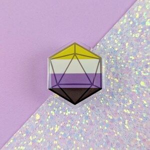 May include: A hexagonal pin featuring a twenty-sided die shape overlaid on a non-binary pride flag. The flag consists of horizontal stripes in yellow, white, purple, and black. The die design is rendered in dark lines over the coloured background.