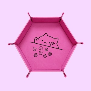 May include: A bright pink, hexagonal dice tray with a black cat illustration peeking over a line, with several dice below. The tray has folded corners and is set against a light pink background.