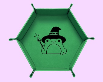 May include: A green, hexagonal dice tray with a black cartoon frog wearing a witch hat and holding a wand. The tray has four corner tabs and is set against a light pink background. The frog is smiling.