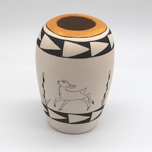 May include: A beige ceramic vase with a black and white geometric pattern around the top and bottom. A running deer and stylized plants are depicted in black. The rim is orange.