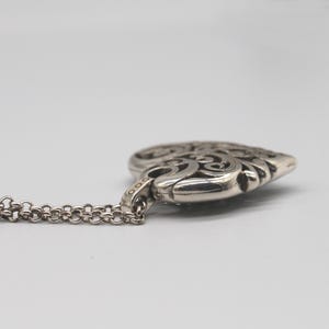 Puede incluir: Un collar con colgante plateado en forma de coraz&oacute;n. El colgante presenta un dise&ntilde;o calado ornamentado con patrones arremolinados. La cadena es de estilo delicado y enlazado. El collar se muestra sobre un fondo liso.