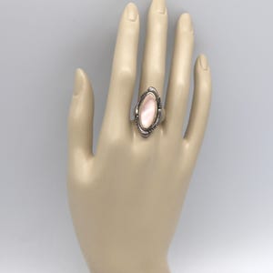 May include: A silver ring with an oval, light pink stone. The ring has a decorative border and is set on a hand. The ring is a piece of jewelry.