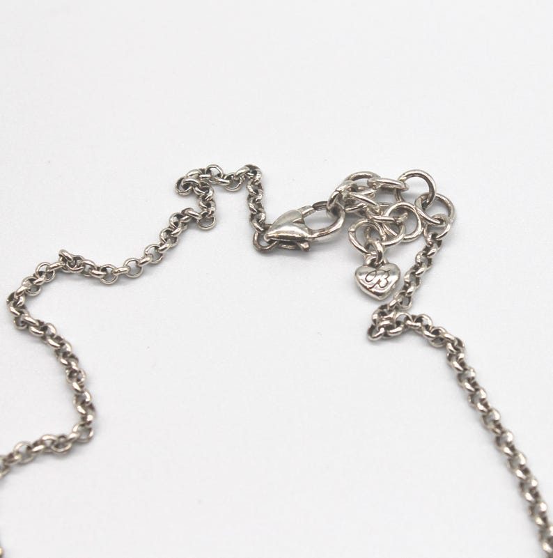 Puede incluir: Primer plano de un collar plateado con una cadena delicada. El collar presenta un cierre de mosquet&oacute;n y un peque&ntilde;o dije en forma de coraz&oacute;n con las iniciales "B3". La cadena es visible sobre un fondo blanco.