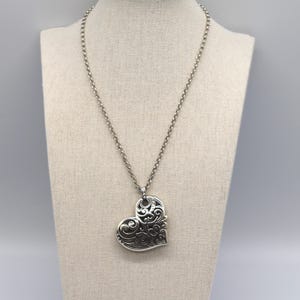 Puede incluir: Un collar con colgante plateado con un gran colgante en forma de coraz&oacute;n calado. El coraz&oacute;n presenta intrincados dise&ntilde;os de espirales y cuelga de una delicada cadena. El collar se exhibe en un maniqu&iacute; de tono neutro.