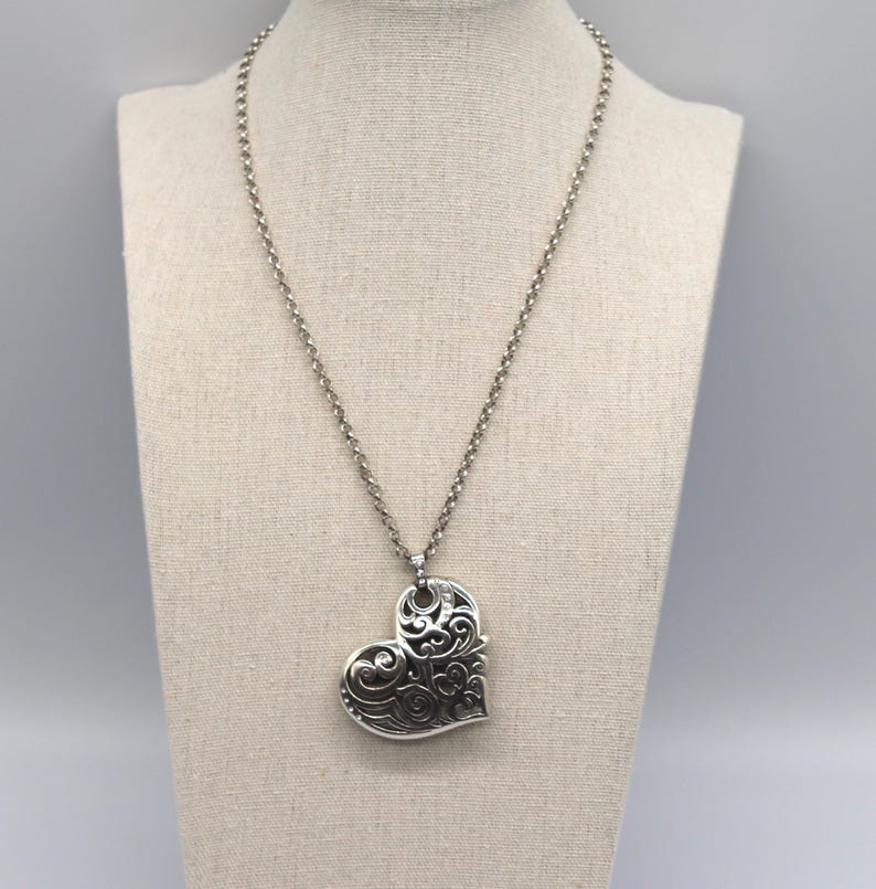 Puede incluir: Un collar con colgante plateado con un gran colgante en forma de coraz&oacute;n calado. El coraz&oacute;n presenta intrincados dise&ntilde;os de espirales y cuelga de una delicada cadena. El collar se exhibe en un maniqu&iacute; de tono neutro.