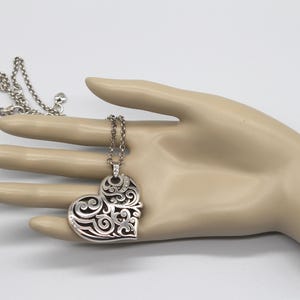 Puede incluir: Collar con colgante de coraz&oacute;n plateado con un dise&ntilde;o intrincado y calado. El colgante en forma de coraz&oacute;n presenta patrones arremolinados y est&aacute; suspendido de una delicada cadena. El collar se muestra sobre un fondo neutro.