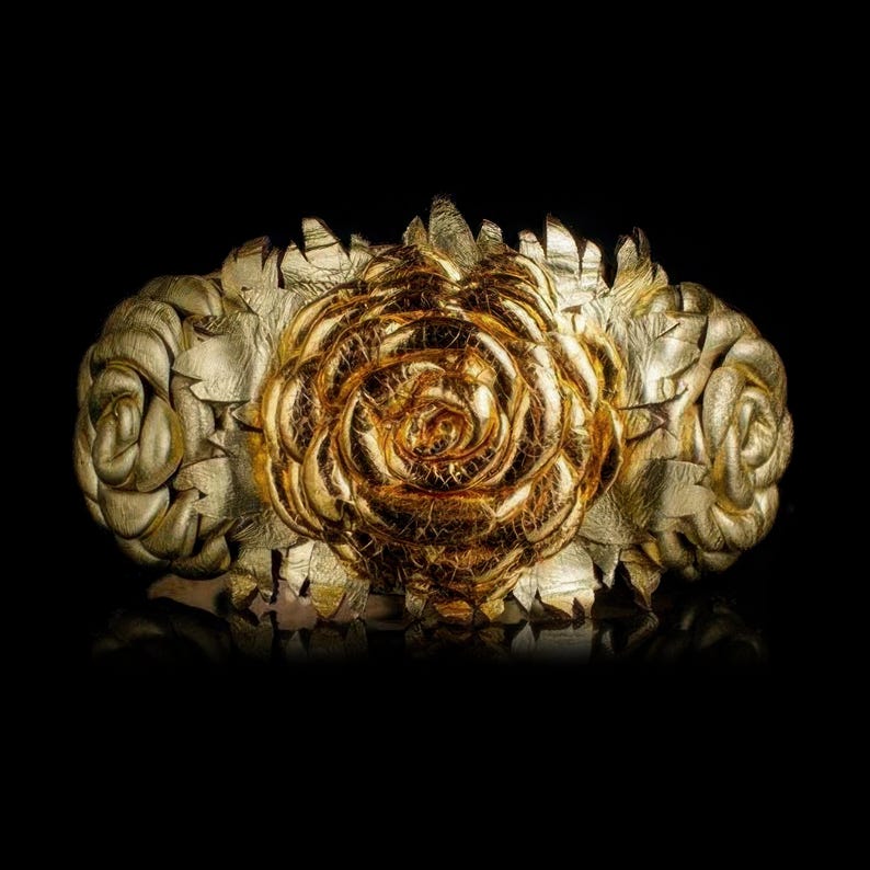 May include: A gold-colored floral crown with a large rose in the center and two smaller roses on either side. The crown is made of what appears to be metal or a similar material, with intricate details and a reflective surface. The background is black.