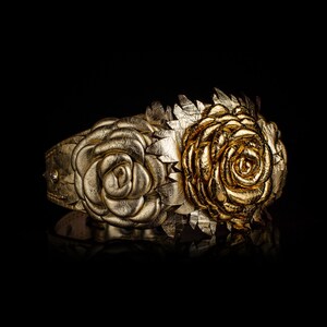 May include: A gold-colored leather cuff bracelet featuring embossed rose and leaf designs. The bracelet has a central rose and two smaller roses on either side, with intricate leaf detailing. The background is black.