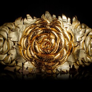 May include: A gold-colored floral crown with a large rose in the center and smaller roses on each side. The crown is made of a metallic material with a textured, crackled finish, and is set against a black background.