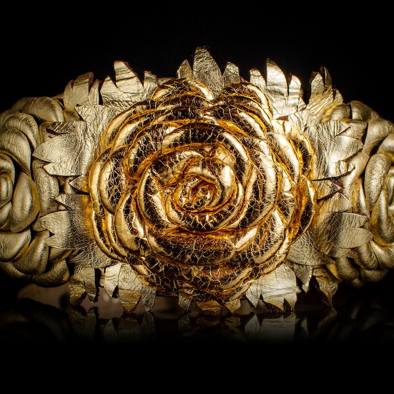 May include: A gold-colored floral crown with a large rose in the center and smaller roses on each side. The crown is made of a metallic material with a textured, crackled finish, and is set against a black background.