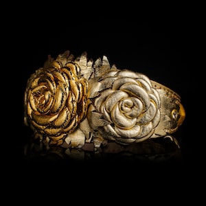 May include: A gold-colored cuff bracelet featuring two large, textured rose designs. The bracelet is made of a metallic material and has a reflective surface. The roses are intricately detailed, with layers of petals. The background is black.