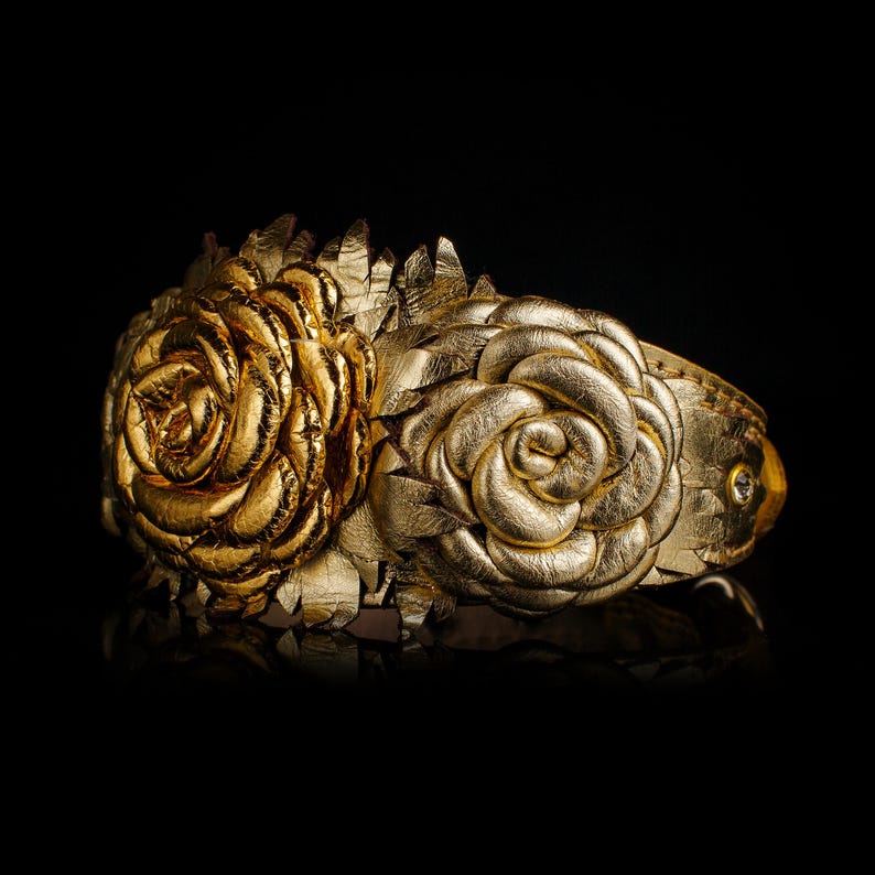 May include: A gold-colored cuff bracelet featuring two large, textured rose designs. The bracelet is made of a metallic material and has a reflective surface. The roses are intricately detailed, with layers of petals. The background is black.