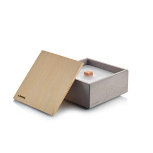 Peut inclure: Un bougeoir carré en béton avec un couvercle en bois. La bougie a une cire blanche et une petite mèche en bois. Le couvercle est en bois clair et porte le mot "abeske" imprimé.