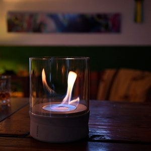 May include: A white and glass tabletop fireplace with a blue flame burning inside. The fireplace is on a wooden table.