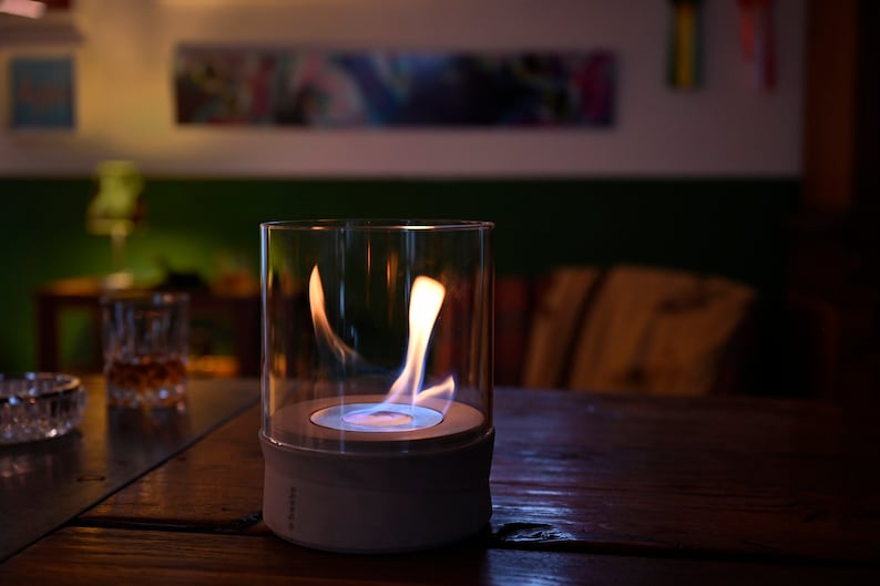May include: A white and glass tabletop fireplace with a blue flame burning inside. The fireplace is on a wooden table.