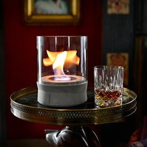 May include: A gray concrete tabletop fire pit with a glass cylinder and a flame burning inside. The fire pit is on a gold-colored table with a glass of whiskey on the table.