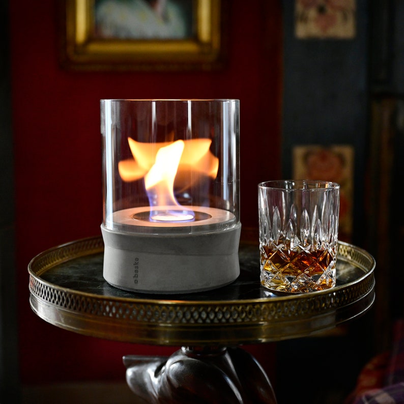 May include: A gray concrete tabletop fire pit with a glass cylinder and a flame burning inside. The fire pit is on a gold-colored table with a glass of whiskey on the table.