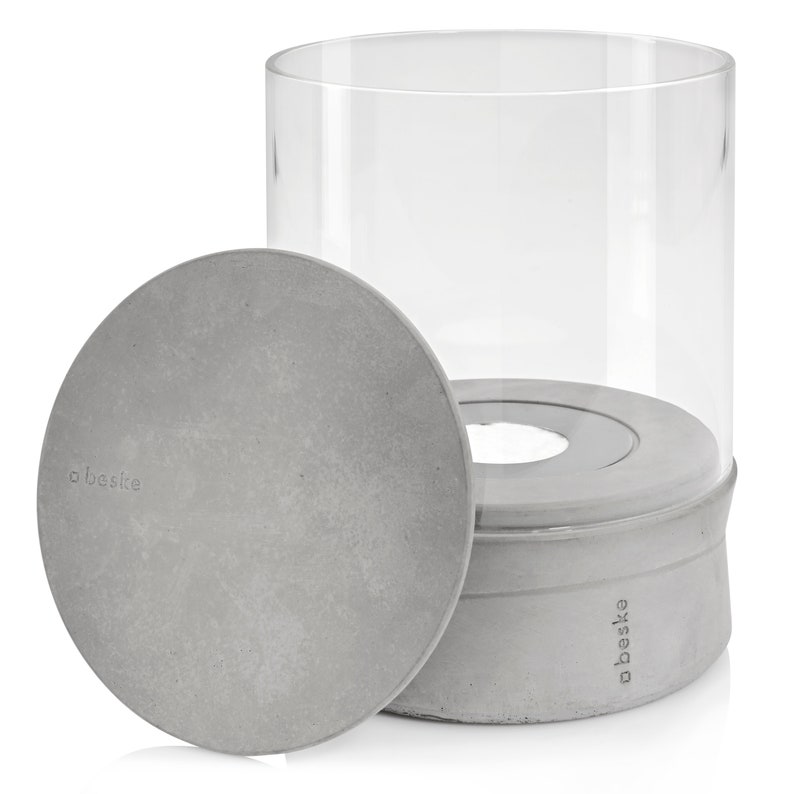 May include: A gray concrete candle lantern with a clear glass cylinder and a removable lid. The lantern has the word "obest&eacute;" printed on the side.
