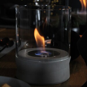 May include: A gray concrete tabletop fireplace with a glass cylinder and a blue flame burning inside.