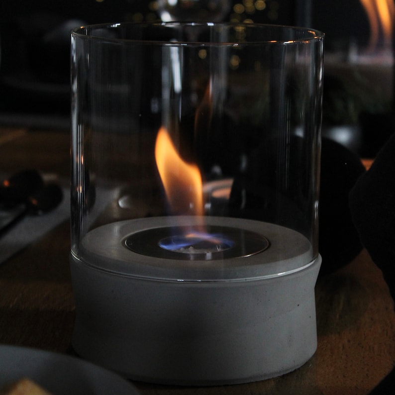 May include: A gray concrete tabletop fireplace with a glass cylinder and a blue flame burning inside.