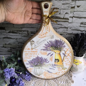 Pu&ograve; includere: Un tagliere in legno bianco e dorato con un design di lavanda. Il tagliere ha un manico ed &egrave; decorato con un motivo floreale e il testo "de Provence".